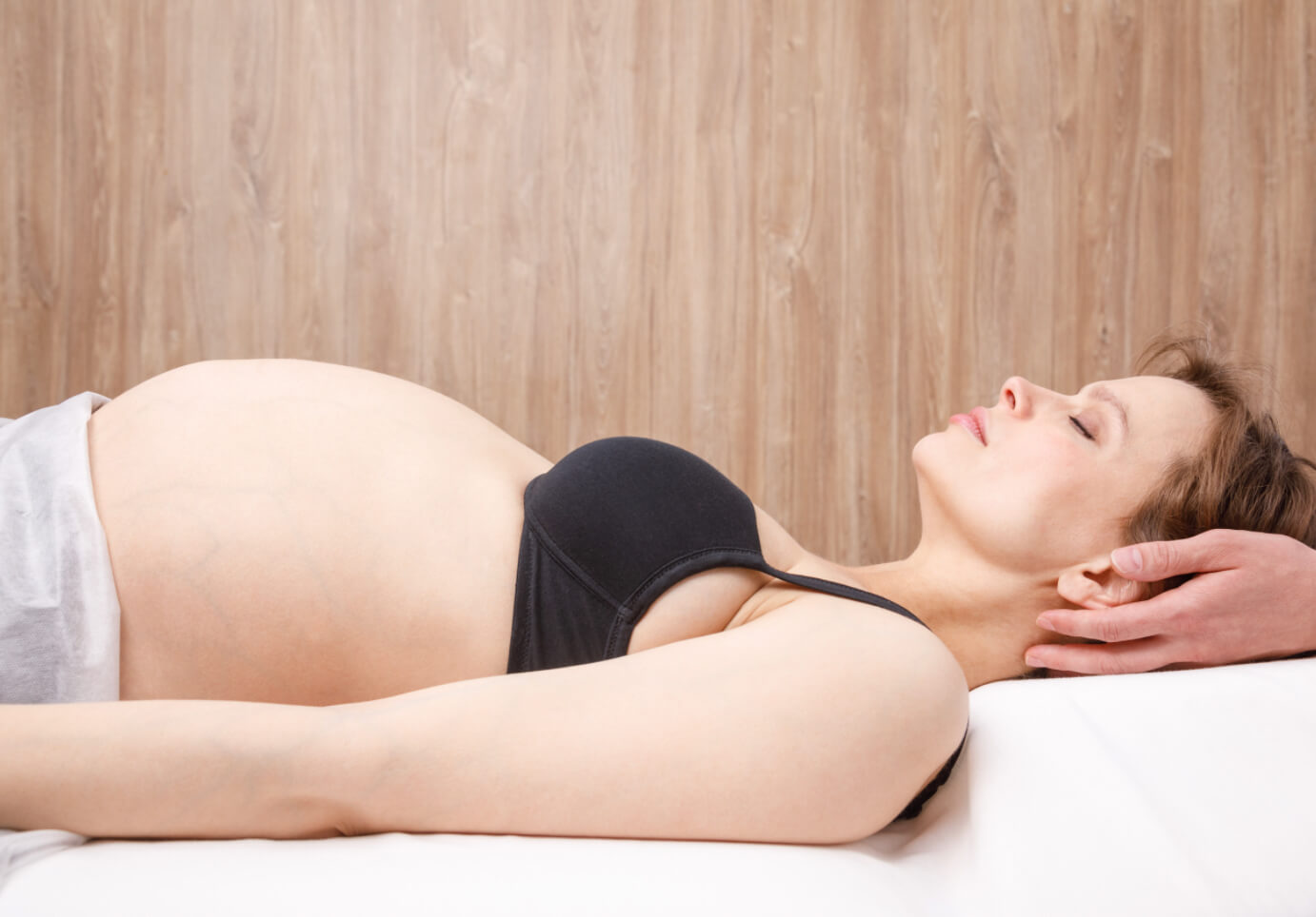 Pregnant white woman in black bra and white sheet with naked belly with neck being treated by white female physio hands 