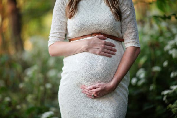 Pregnant woman standing in nature in a white dress holding her belly.